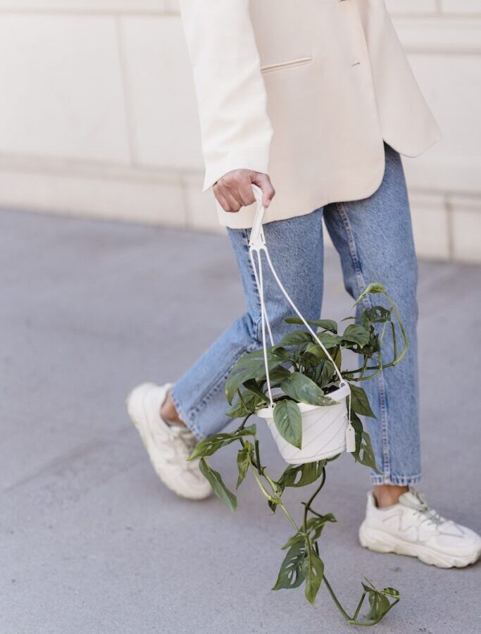 Crop anonymous female in stylish outfit and sneakers walking on sidewalk with hanging planter with green foliage on street in city