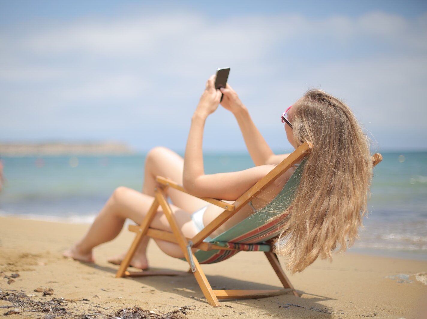 Woman in Blue and White Bikini Sitting on Brown Wooden Chair on Beach While Using Smartphone