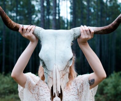 woman holding white and black animal skull wearing white lace floral sleeveless dress near green leaf trees