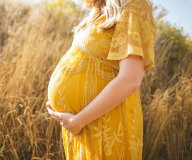 pregnant woman wearing yellow floral dress standing while touching her tummy and facing her right side near brown field during daytime