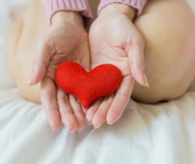 Unrecognizable female sitting with bare legs on white sheet with small red heart in hands in light room in daytime