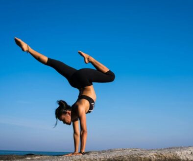 A woman performing a yoga handstand on a rock by the sea under a clear blue sky.