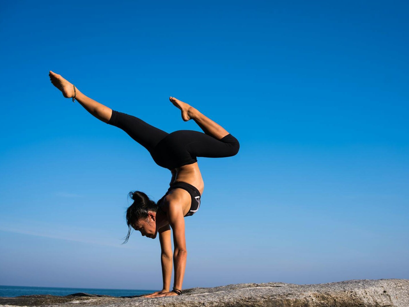 A woman performing a yoga handstand on a rock by the sea under a clear blue sky.