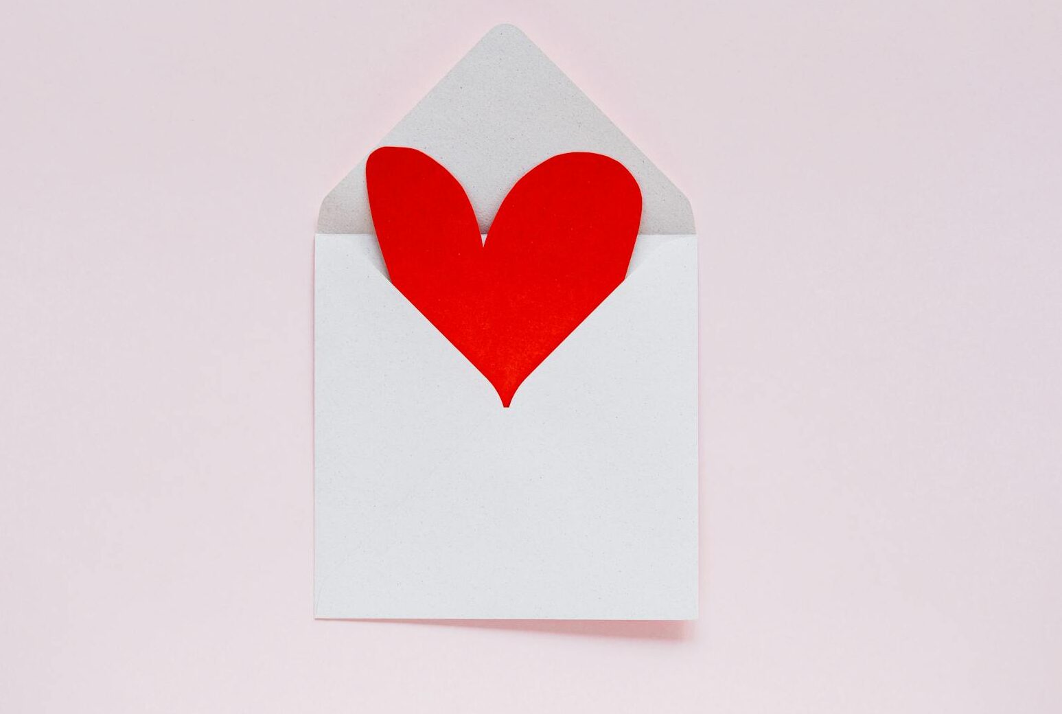 A red heart paper cutout in an envelope on a pastel pink background, ideal for Valentine themes.