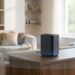Black rectangular speaker on a wooden coffee table in a bright living room with beige seating and plants nearby, sunlit scene