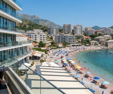 Couple dining on a hotel balcony overlooking a crowded sandy beach and blue-green bay with boats anchored offshore, mountains in the background.