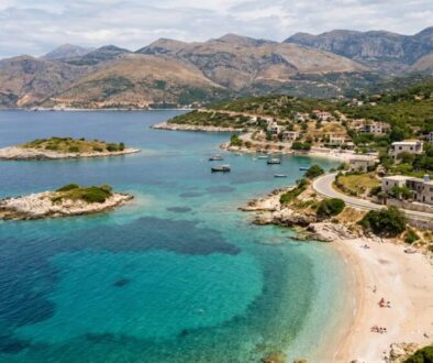 Aerial view of a turquoise bay with a sandy beach, rocky islets, and a hillside village along a curving road.