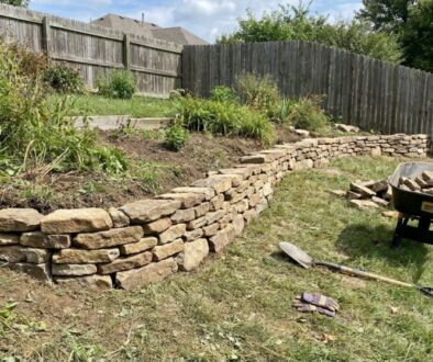 Curved stacked-stone retaining wall being built in a backyard, with a wheelbarrow full of rocks and tools on the grass nearby.