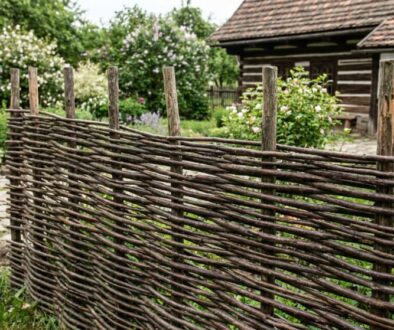 A rustic woven wooden fence enclosing a garden with flowering bushes and a timber house in the background
