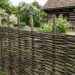 A rustic woven wooden fence enclosing a garden with flowering bushes and a timber house in the background