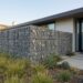 Modern house entry with a tall gabion stone wall, concrete stepping stones, and drought-tolerant plants in the front yard.