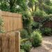 Backyard garden with wooden privacy fences, raised planter boxes, and a gravel path lined by assorted shrubs and herbs.