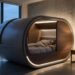 Futuristic oval bedroom pod with a bed inside, warm lighting, beside a wooden nightstand and lamp in a high-rise room overlooking a city skyline.