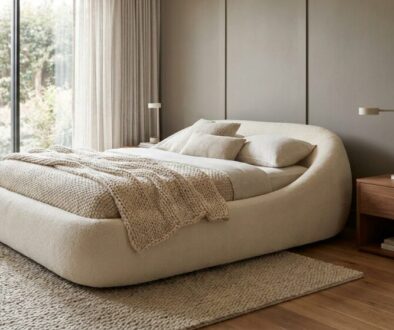Curved cream upholstered bed with a chunky knit throw in a neutral bedroom, beside a wooden nightstand with a lamp and plant.