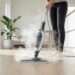 Person using a steam mop to clean a bright wooden floor in a modern kitchen/living space, steam rising from the mop head.