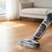 Person using a cordless stick vacuum to clean a wooden floor in a bright living room with a sofa in the background.