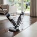 Person using a cordless vacuum to clean a wet spill on the wooden floor in a bright living room, near a sofa.