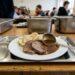 Plate with sliced roast beef, gravy, and bread on a metal cafeteria tray in a busy dining hall.