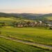 Sunset over rolling green fields with a small village of red-roofed houses and a winding river nearby.