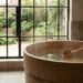 Indoor wooden soaking tub with steam; a hand uses a wooden ladle to stir the water, next to a towel and mug by a bench, with large glass doors opening to a garden.