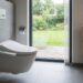 Modern bathroom with a wall-mounted smart toilet and bidet controls, opening to a green garden beyond a glass door.