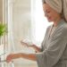Smiling woman with a towel on her head and a gray robe, washing foamy soap from her hands at a bright bathroom sink.