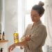 Woman in a beige bathrobe applies oil to her forearm in a bright bathroom; bottles of oil line a wooden shelf nearby.
