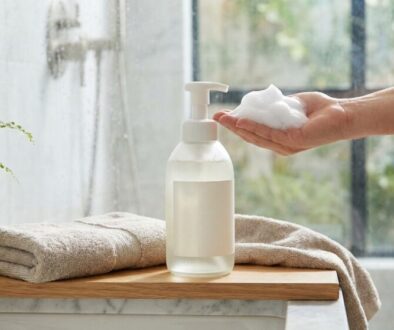 Hand holding a small amount of white foam soap above a pump bottle on a wooden tray, with towels and a potted plant nearby in a bathroom setting.