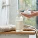 Hand holding a small amount of white foam soap above a pump bottle on a wooden tray, with towels and a potted plant nearby in a bathroom setting.