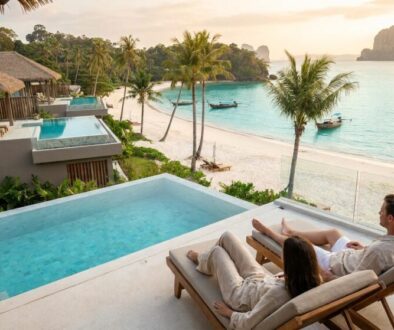 Couple lounging on wooden sunbeds beside an infinity pool, overlooking a tropical beach and turquoise bay at sunset.
