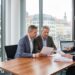 Three professionals in a meeting at a wood table, reviewing documents with a laptop showing charts, in a bright office with city buildings outside.