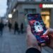 Close-up of a smartphone showing a cracked padlock graphic, symbolizing a cybersecurity breach in a city street with a bank in the background, blurred.