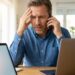 Man at a desk with two laptops, holding a phone to his ear and looking stressed as a red alert icon appears on the left screen.