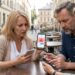Three adults at an outdoor cafe examining smartphones, the woman showing her phone with a red alert icon on screen.