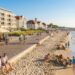 Beachfront promenade with cafes and outdoor dining, cyclists on the boardwalk, and families playing on the sandy shore.