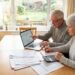 Older couple at a wooden dining table reviewing documents with a laptop and calculator, indoors by a large garden window.