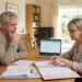 Older man and woman sit at a dining table reviewing documents with a laptop open in a bright living room.