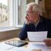 Older man sits at a kitchen table by a window, studying paper documents with a tablet nearby and thinkingowy hand on his chin.