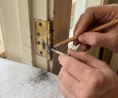Hands filing a brass door hinge on a light door frame, pencil in hand nearby on a work surface.