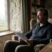 Older man with a beard sits in a cozy armchair by a stone wall window, reading papers in a rural study.