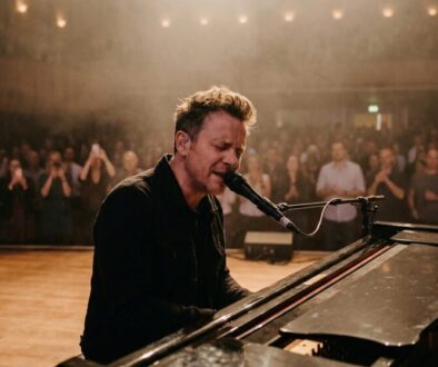 Male musician at a grand piano singing into a microphone on stage as a crowd watches in a dimly lit venue