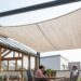 Rooftop terrace with a beige fabric shade sail over a wooden dining table where three people chat, with potted plants nearby.