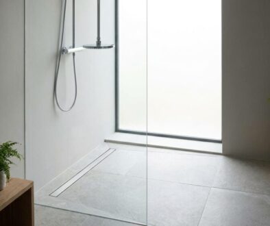 Minimalist bathroom with a glass shower enclosure, large window, gray tile floor, and two wooden benches with potted ferns.