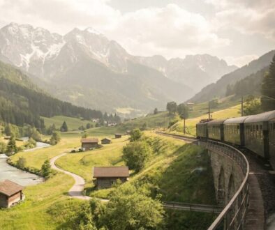 Steam line train curves along a stone viaduct through a green valley with a river and snow-capped mountains in the distance