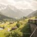 Steam line train curves along a stone viaduct through a green valley with a river and snow-capped mountains in the distance