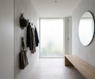 Minimalist entryway with wall hooks holding coats, hats, and bags, a frosted window at the end, and a round wall mirror opposite a wooden bench