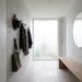 Minimalist entryway with wall hooks holding coats, hats, and bags, a frosted window at the end, and a round wall mirror opposite a wooden bench