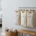 Laundry sorting system with labeled fabric bags (DARKS, LIGHTS, DELICATES) hanging on a wall rod in a bright bedroom.