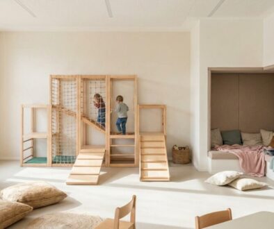 Bright children's playroom with a wooden climbing structure, ramps, and a cozy reading nook by a windowed wall shelves filled with books and cushions nearby.