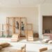 Bright children's playroom with a wooden climbing structure, ramps, and a cozy reading nook by a windowed wall shelves filled with books and cushions nearby.